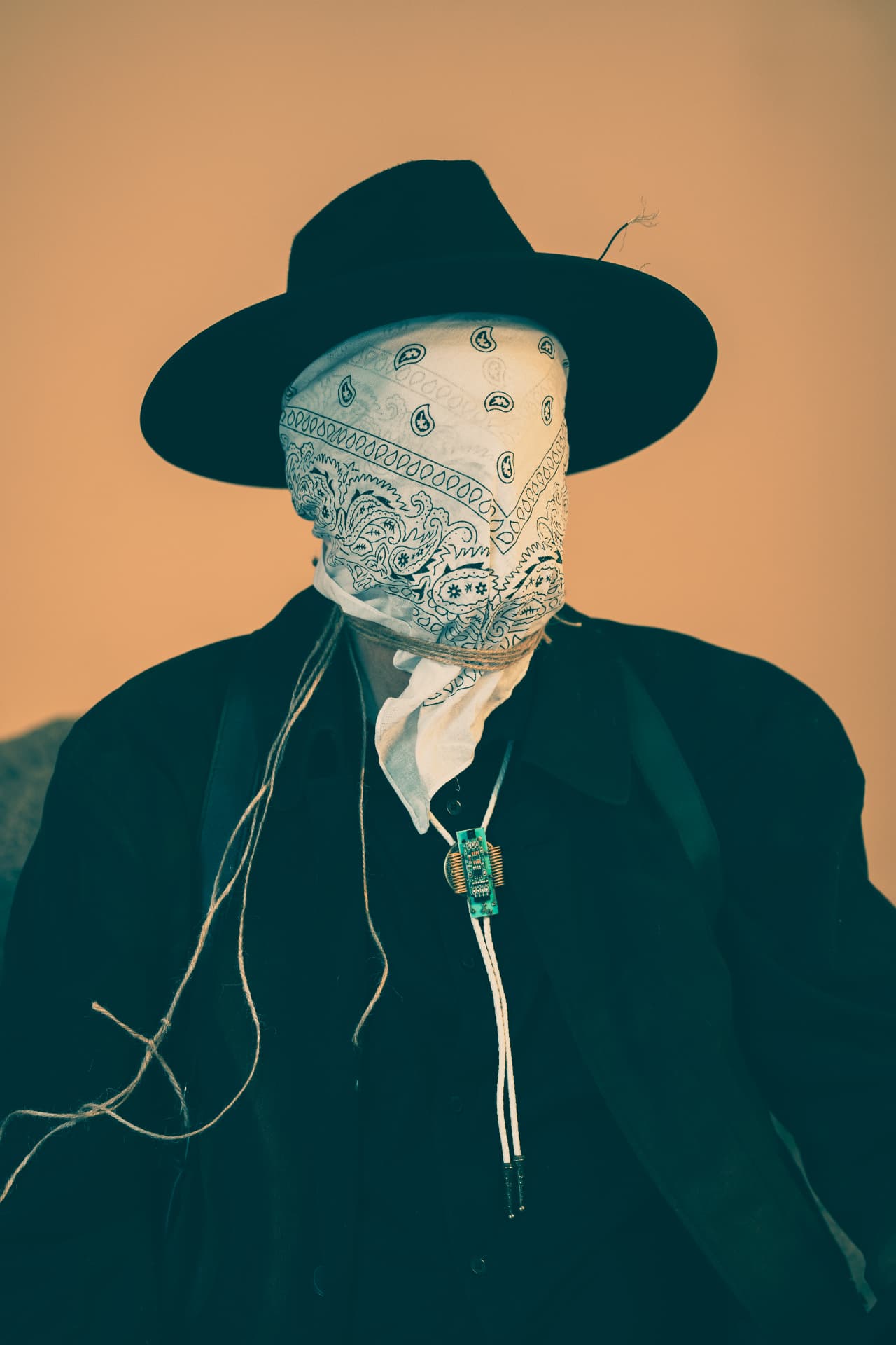 The Traveler in a black hat, a white bandana covering his face, against a warm tan background