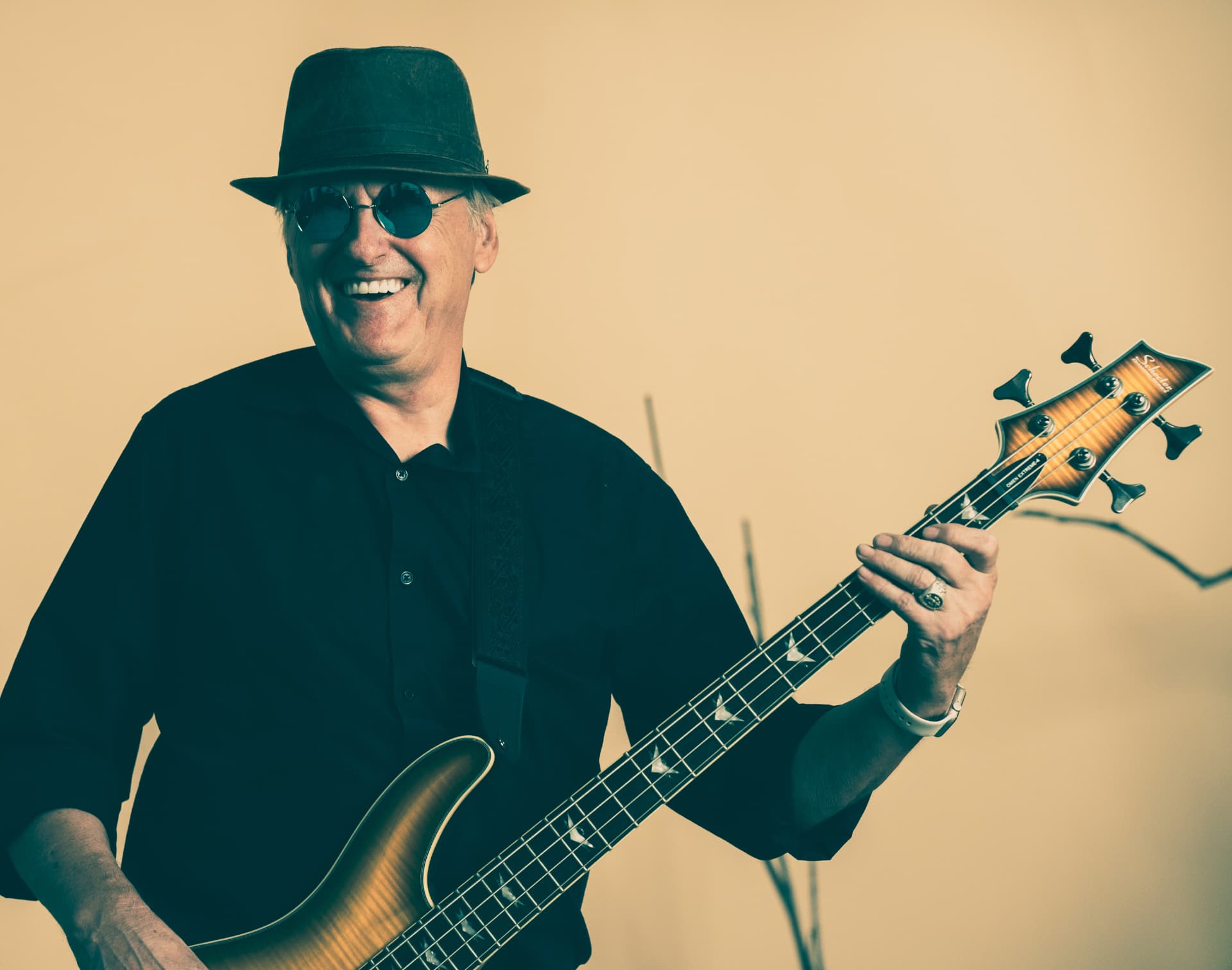Chuck Wooke smiling in a dark hat, round blue sunglasses and black shirt, playing a sunburst bass guitar