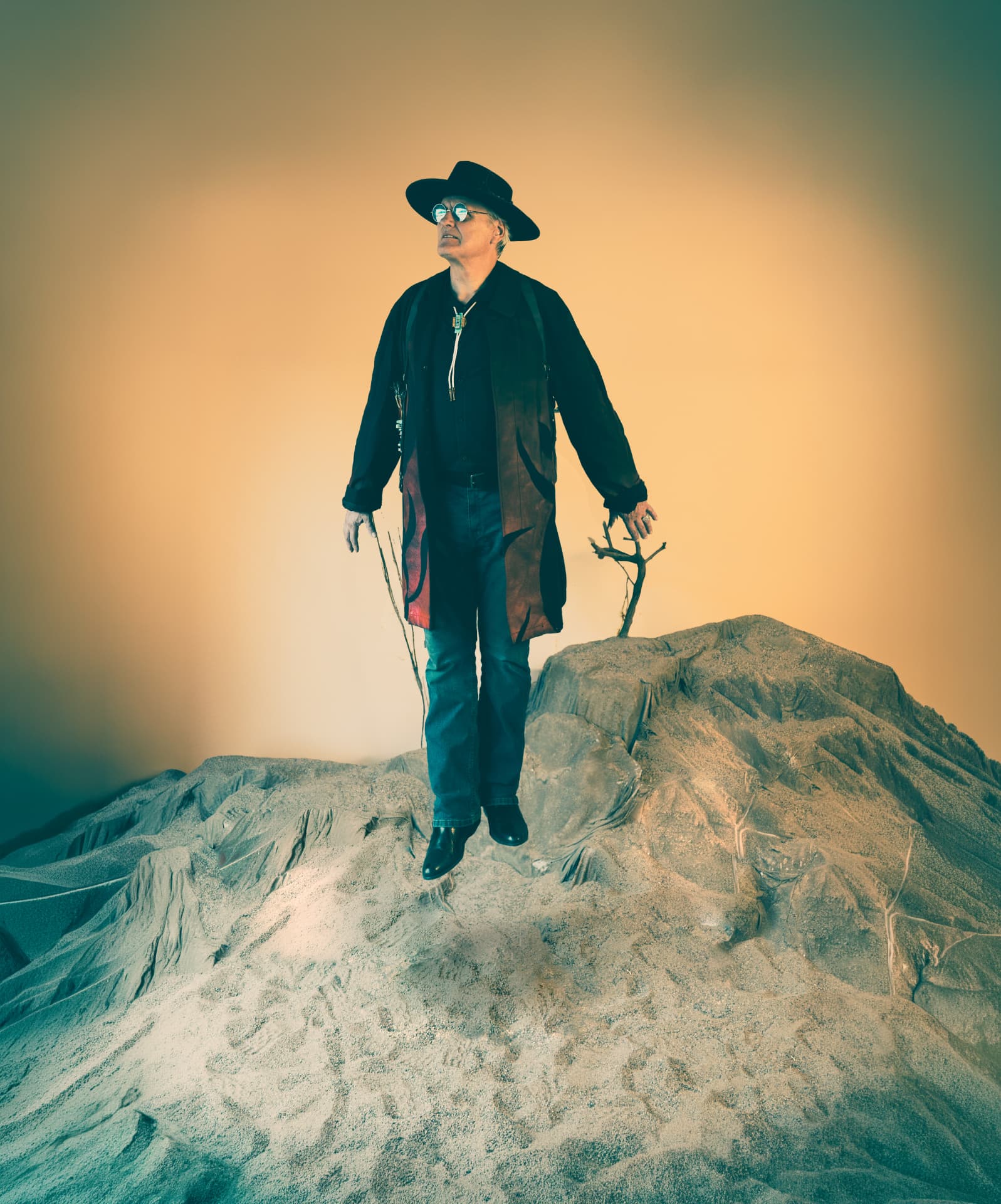 Chuck Wooke as The Traveler, in black hat and long coat, suspended above a sculpted desert mound against a glowing amber sky